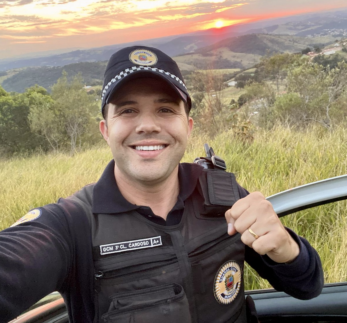 Lucas Cardoso com a farda da Guarda Civil Municipal, junto a uma viatura, com paisagem ao pôr do sol