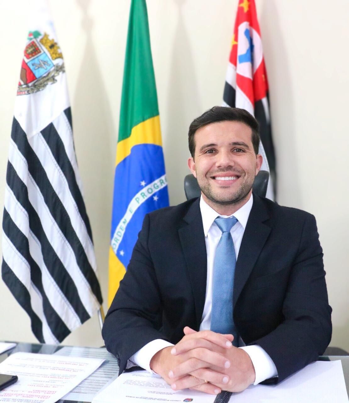 Lucas Cardoso sentado à mesa, de terno e gravata, com bandeiras ao fundo